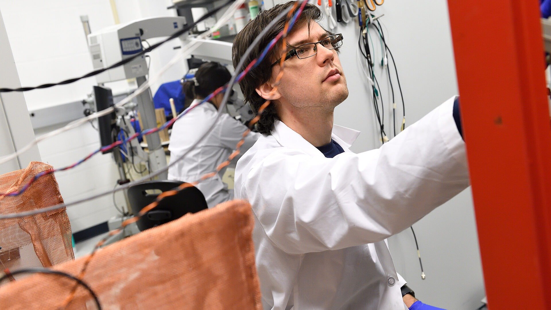 A male graduate student works in the lab amongst lots of wires.