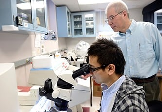 A student looks into a microscope as a faculty member stands behind him.