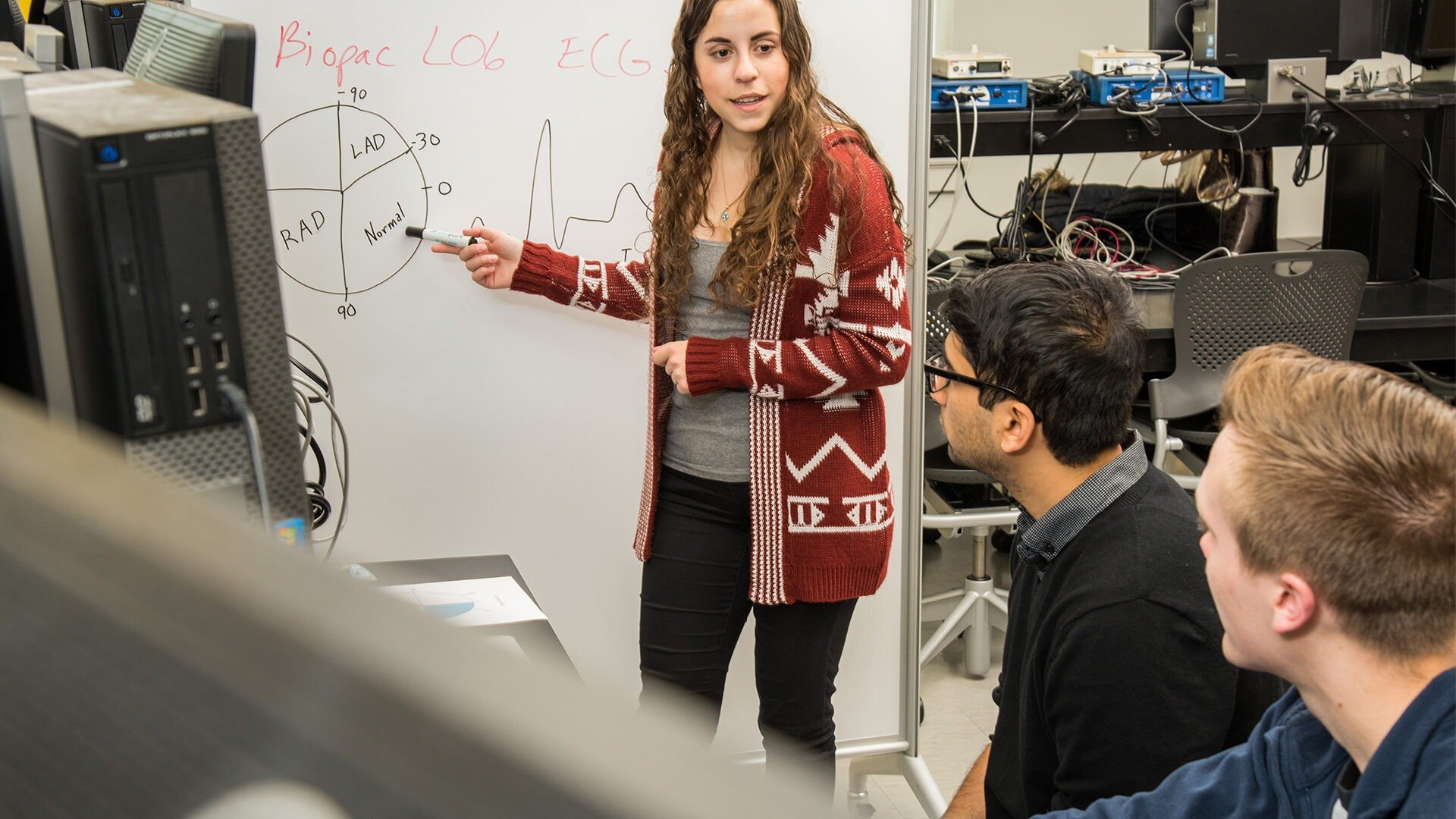 A female student stands at a whiteboard discussing something with two classmates.