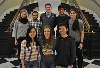 A group of students stand in front of a curved staircase and smile at the camera.