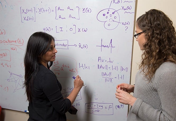A faculty member and a student discuss equations at a white board.