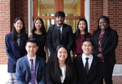 A group of well-dressed students pose in front of a set of glass doors and a brick wall.