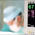 A clinicial is working in the background and a patient monitor is in the foreground.