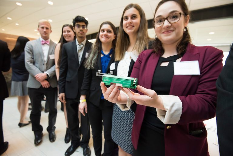 A team of students are dressed up for Design Day and one is holding a prototype.