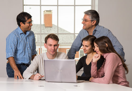 A group of students and faculty chat while working around a laptop.