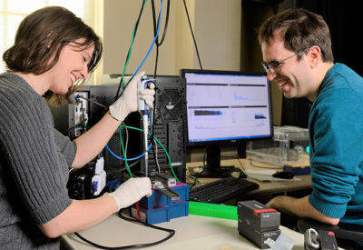 Two faculty members are working at a desk with a computer and pipettes.
