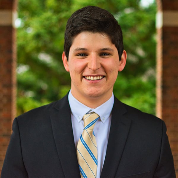 Santiago Balza is wearing a dark suit and yellow tie, smiling for the camera.