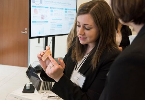 A female student in a black blazer holds up a small prototype as she talks with another person.