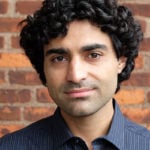 Reza Kalhor is standing in front of a brick wall wearing a blue button down shirt.