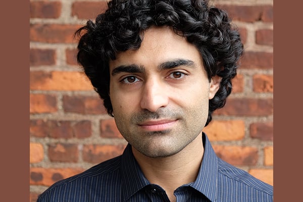 Reza Kalhor is standing in front of a brick wall wearing a blue button down shirt.