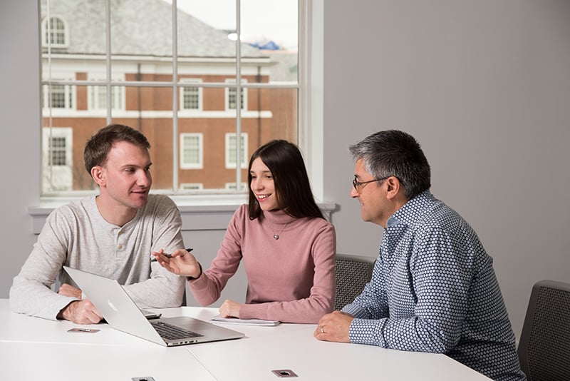 Two students sit with a professor at a table with a laptop.