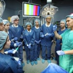 BME students are dressed in scrubs and listening to a surgeon in the OR.