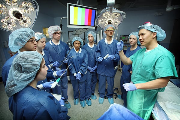 BME students are dressed in scrubs and listening to a surgeon in the OR.