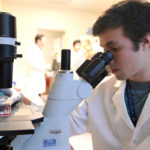 An student is wearing a white coat while he looks into a microscope.