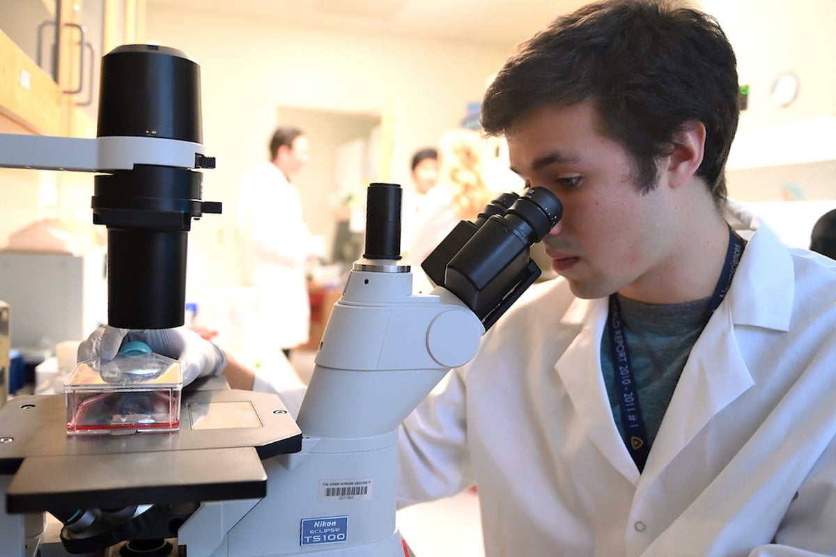 An student is wearing a white coat while he looks into a microscope.