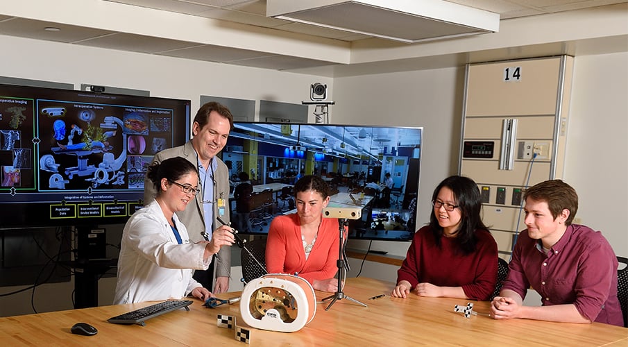 Three students sit around a table with a male faculty member and a female clinician and discuss the model on the table.