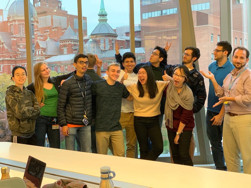 Josh Doloff and the students in his lab gather for a fun group photo in front of windows looking out onto the School of Medicine campus.