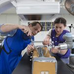 A female student works with a faculty member on a model spine under a medical imaging device.