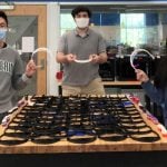 Three students are standing next to a table filled with 3-D printed visors.