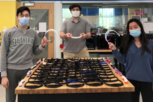 Three students are standing next to a table filled with 3-D printed visors.