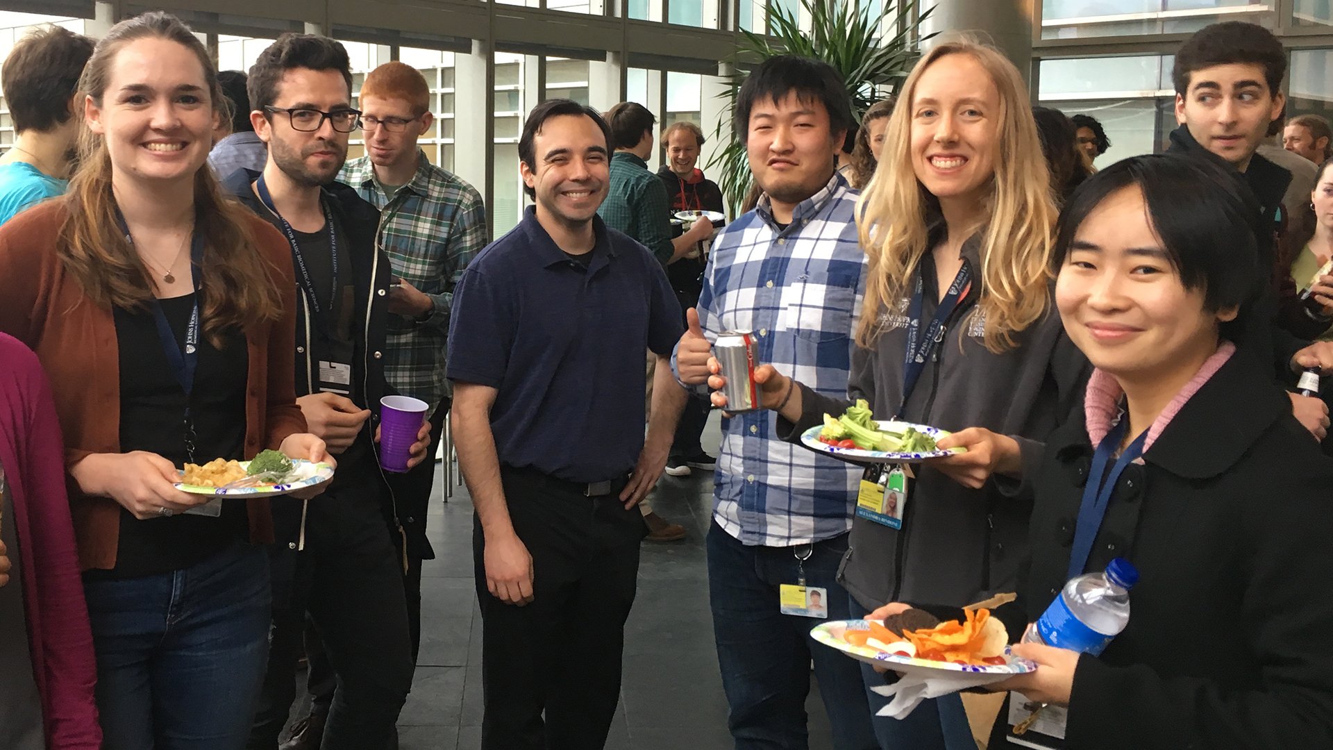 A group of graduate students are holding food and drinks and enjoying a social hour.