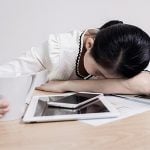 A woman is resting her head on her desk with a cup of coffee in her hand.