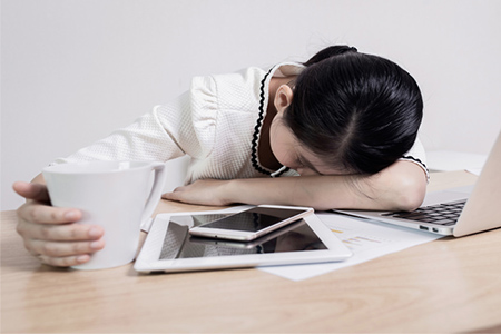 A woman is resting her head on her desk with a cup of coffee in her hand.