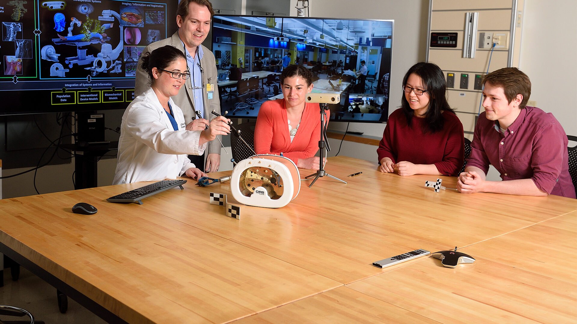 A group of students sit at a table watching a professor and clinician teach a lesson.