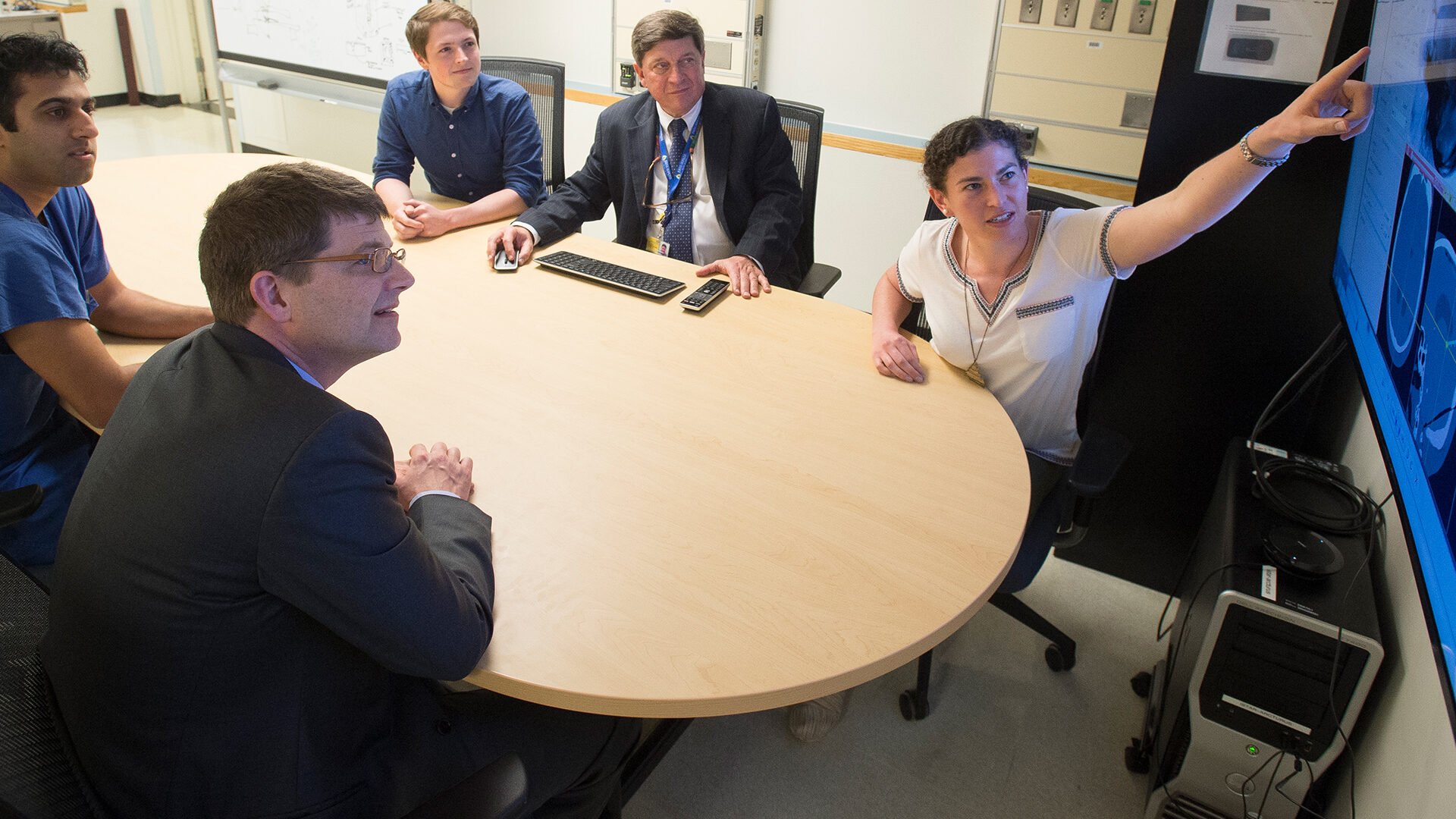 A group of people sit around a table while a woman presents information on a large monitor.