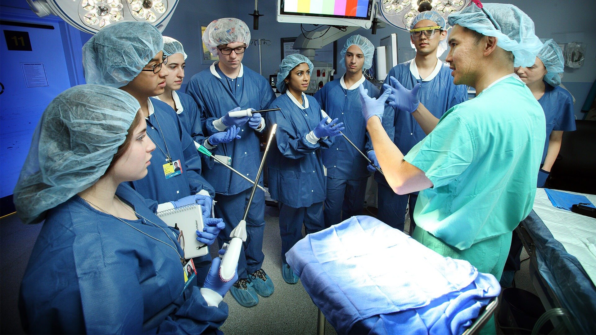 A group of students are dressed in scrubs in an OR room listening to a surgeon.