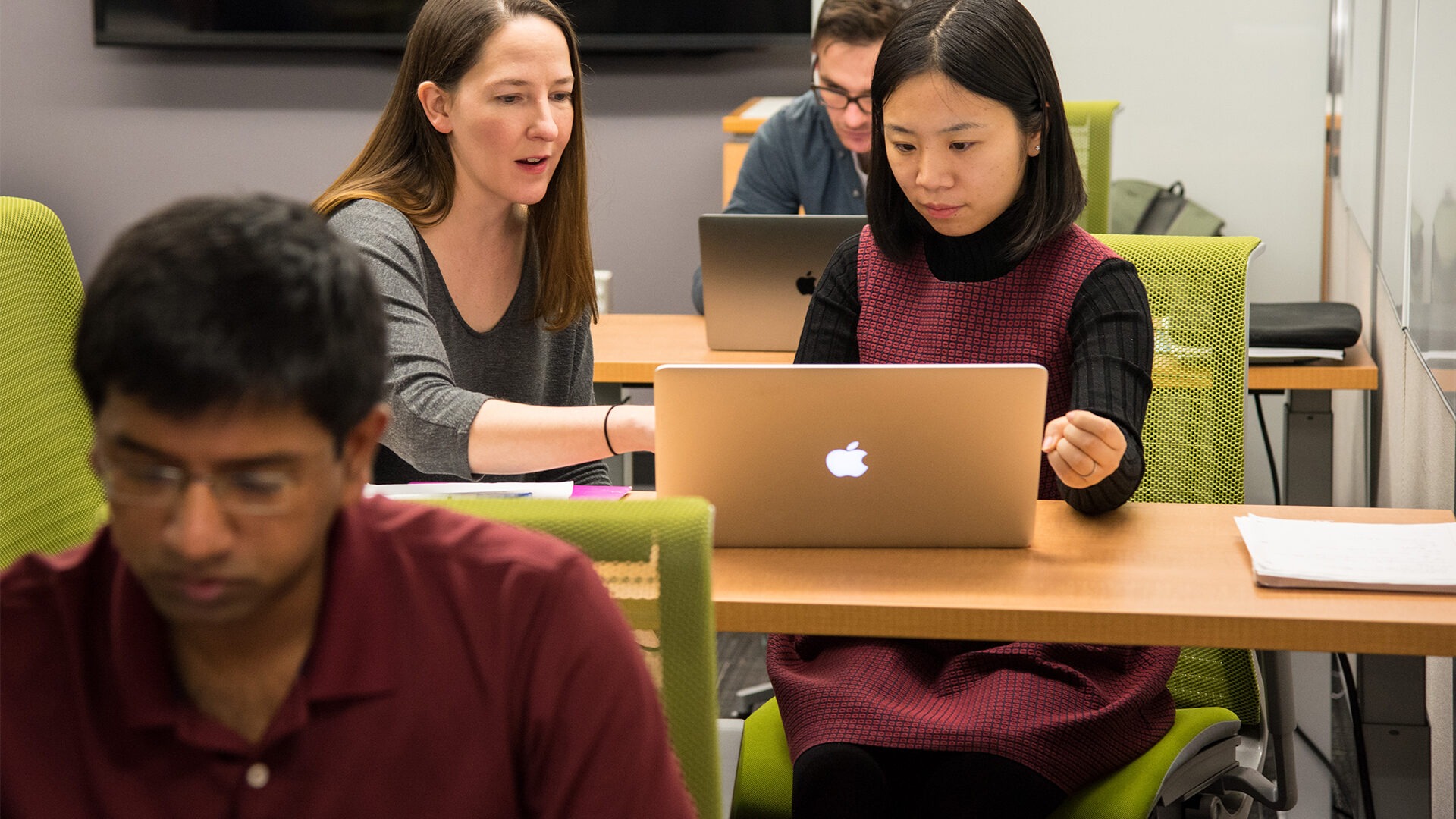 A female professor is working with a female student at a desk with other students in the background.