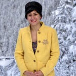 Alaleh Azhir is dressed in a yellow coat and black hat, and is standing in front of snow-covered trees.