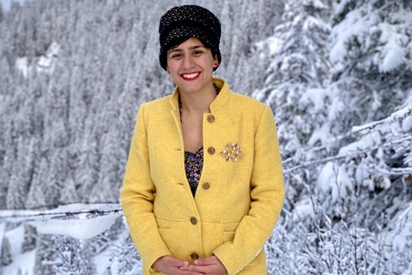 Alaleh Azhir is dressed in a yellow coat and black hat, and is standing in front of snow-covered trees.