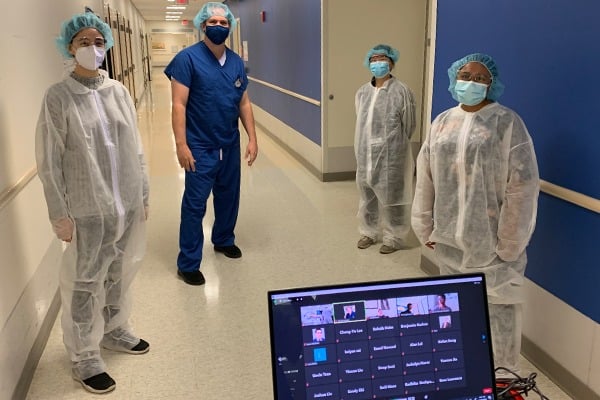 Three students and one professor are wearing scrubs in the hall of a hospital with a computer showing more students on a Zoom meeting.