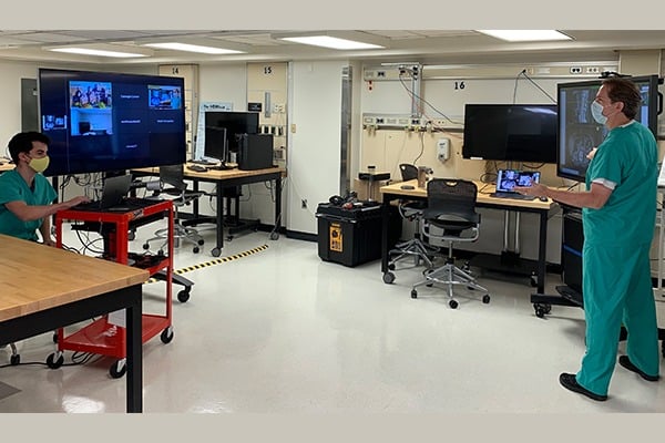 A professor in scrubs and a mask is teaching to a virtual classroom over Zoom.