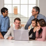 A group of male and female students gather around a laptop with a male professor.