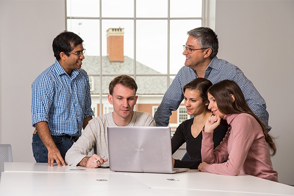 A group of male and female students gather around a laptop with a male professor.