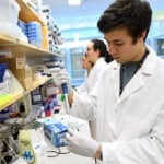 A male student is in a wet lab wearing a white coat and holding a pippette.