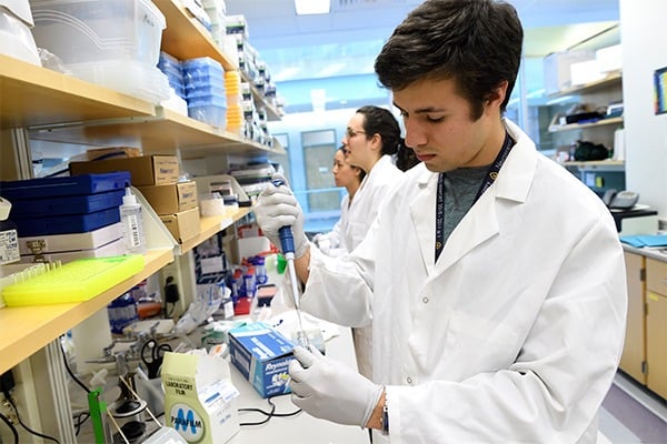 A male student is in a wet lab wearing a white coat and holding a pippette.