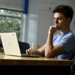 A student sits at a table with his laptop open in front of him.