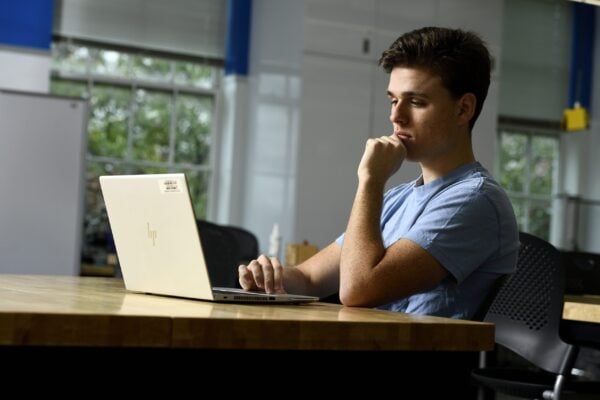 A student sits at a table with his laptop open in front of him.