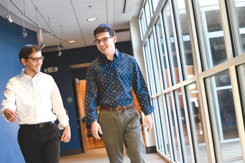 Two males students are walking down a hall with windows.