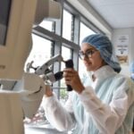 A female student is working with a large piece of medical machinery and wearing scrubs and a cap.