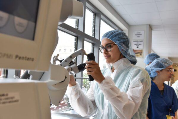 A female student is working with a large piece of medical machinery and wearing scrubs and a cap.