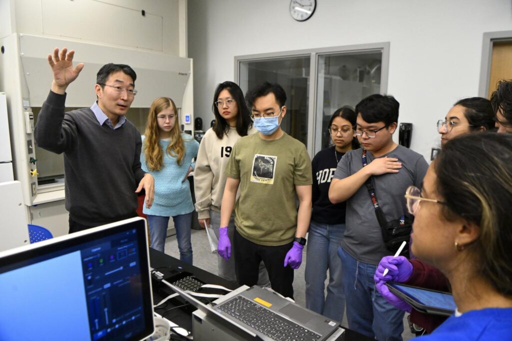 A group of students stand around a professor teaching a class.