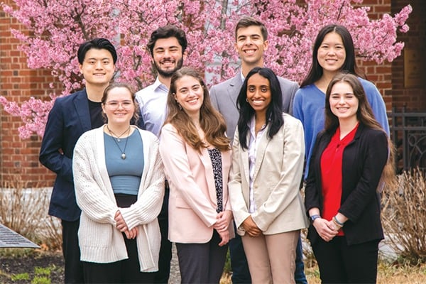 A group of students stands together for a photo in front of a flowering bush.