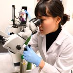 A woman is wearing a lab coat and looking into a microscope.