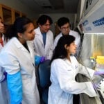 A group of students watch over the shoulder of a female professor working under a hood.