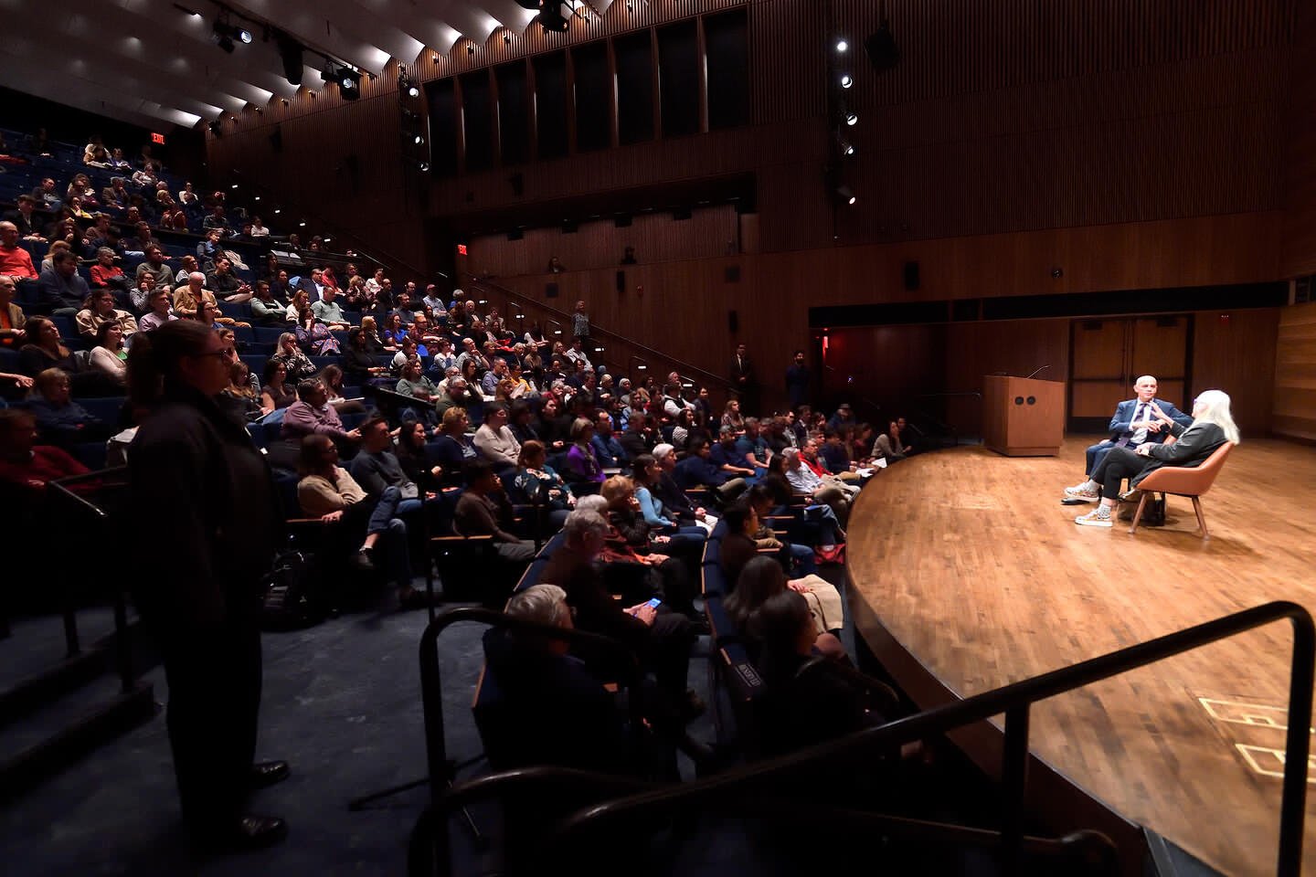 Man and woman talk on a stage in front of an audience.
