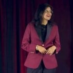 A portrait of Dr. Annie Kathuria, standing on stage with a red velvet curtain background, wearing a burgundy blazer with black lapels and glasses, smiling while looking to the right.